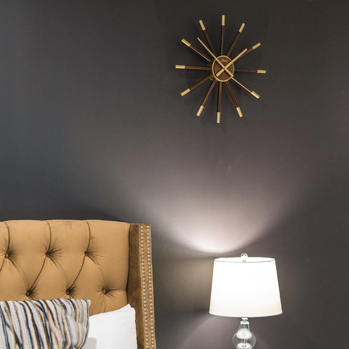 Sunburst style wooden clock hanging above bed and lamp on dark grey wall.