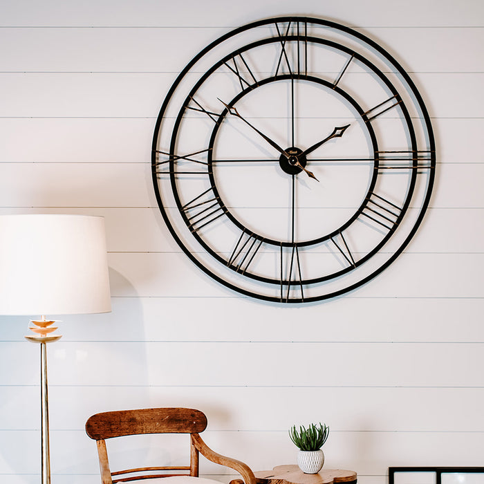Large decorative wall clock with Roman numerals on a white wall above chair and lamp