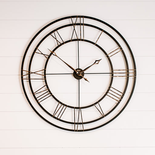 Large round wall clock with black frame and Roman numerals on a white wooden background