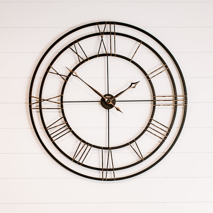 Large round wall clock with black frame and Roman numerals on a white wooden background