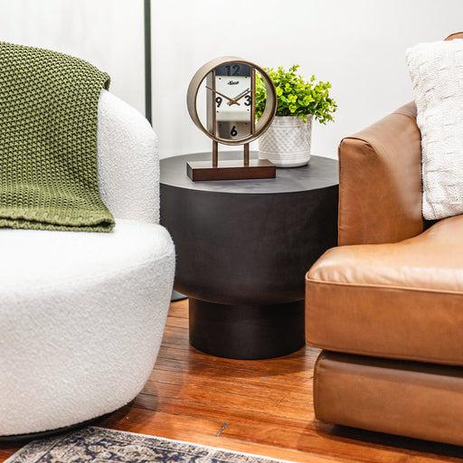 Decorative clock with mirrored face on side table next to green plant flanked by armchairs. 