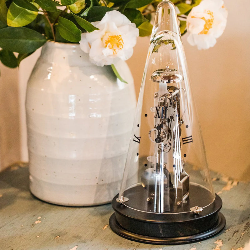 Decorative clock under a glass dome on a wooden table with a vase and flowers in the background