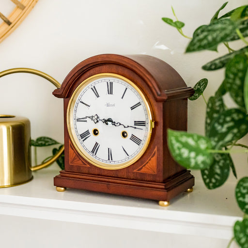 Wooden mantel clock on white shelf with green plant alongside.