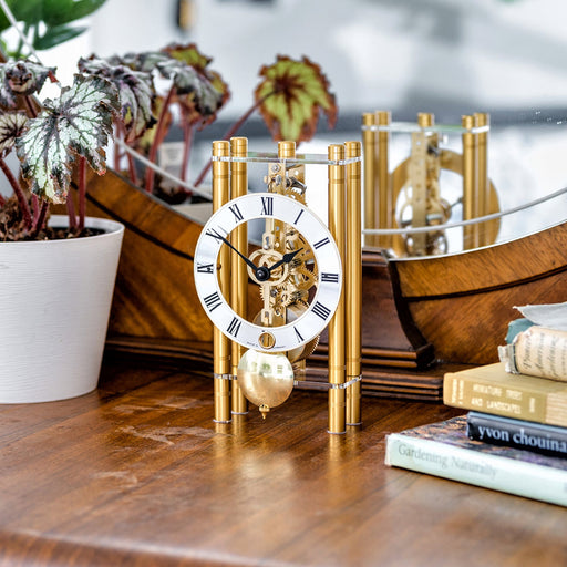 Gold mechanical clock sitting on wooden dresser next to books and potted plant
