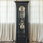 Black grandfather clock with brass and brushed nickel highlights against a white curtain background