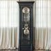 Black grandfather clock with brass and brushed nickel highlights against a white curtain background