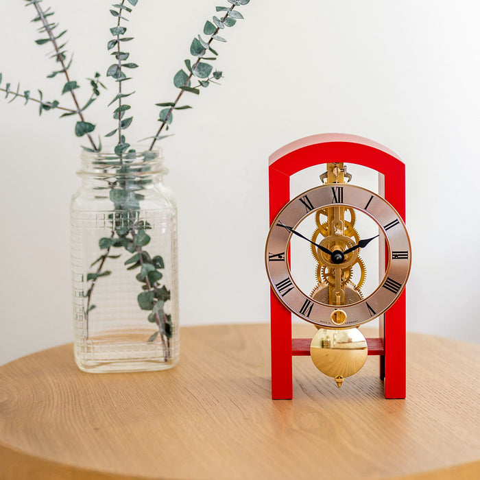 Small red clock with brass mechanism on a wooden surface next to a clear vase with greenery.