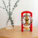 Small red clock with brass mechanism on a wooden surface next to a clear vase with greenery.