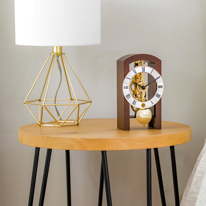 Walnut clock with brass gears and silver face on wooden side table with a gold geometric lamp 