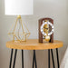 Walnut clock with brass gears and silver face on wooden side table with a gold geometric lamp 