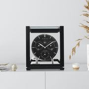Black perpetual calendar clock with black face on white cabinet with books and tan plants on the side.