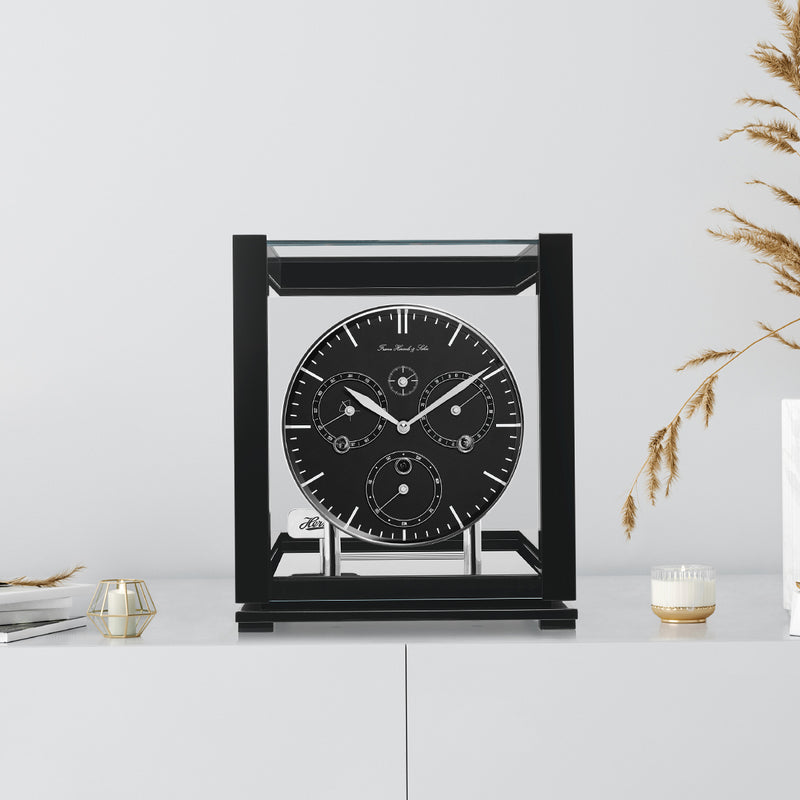 Black perpetual calendar clock with black face on white cabinet with books and tan plants on the side.