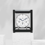 Black perpetual calendar clock with white face on white marble pedestal in white room.