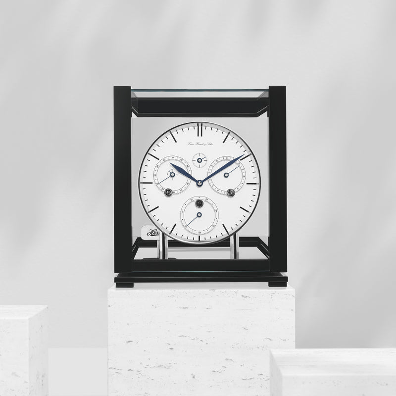 Black perpetual calendar clock with white face on white marble pedestal in white room.