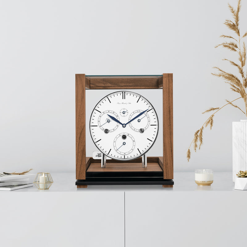 Walnut perpetual calendar clock with white face on white cabinet with books and tan plants on the side.