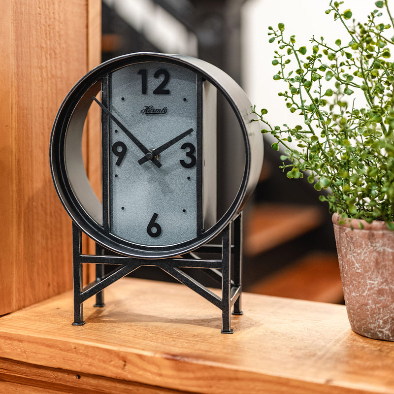 Vintage style metal clock on wooden shelf next to green plant. 