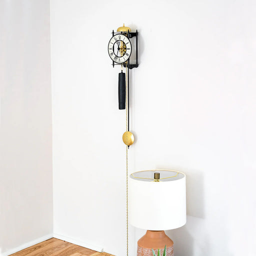 Black skeleton clock with brass pendulum hanging on white wall above decorative lamp