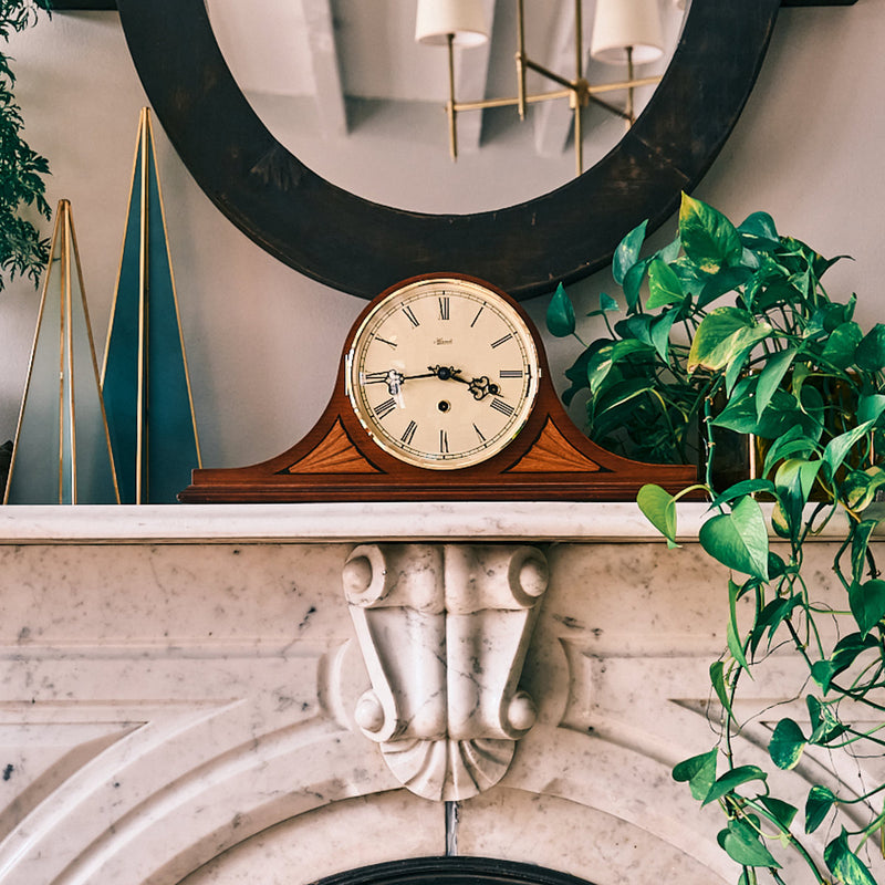 Vintage style tambour clock on marble mantel next to green plant and decorations.