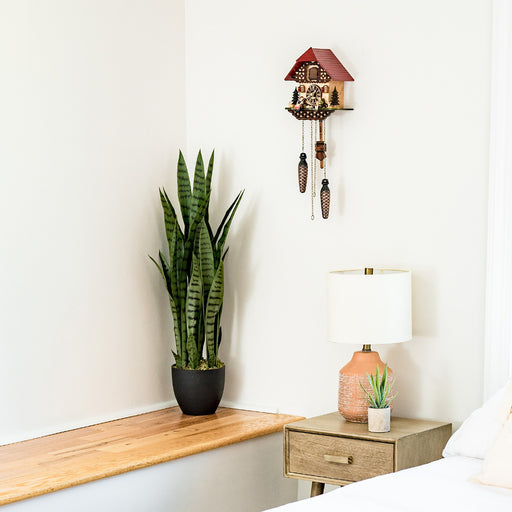 Cuckoo clock with red roof and decorative design hanging on white wall above green plant and lamp on nightstand