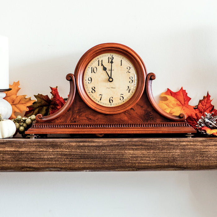 Cherry wood mantel clock on wooden shelf surrounded by fall leaves on white wall.