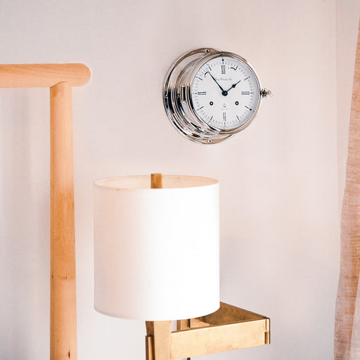 Silver ship's bell clock hanging on white wall next to a wooden bed and beige lamp.