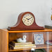 Wooden mantel clock on glass-fronted shelf in front of white wall.