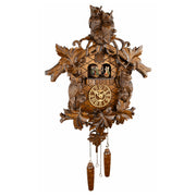 An ornate hand-carved wooden cuckoo clock featuring two owls at the top, leaf and bird motifs, a central clock face with Roman numerals, small figurines in the window, and two dangling wooden weights below, shown against a plain white background.