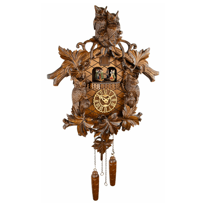 An ornate hand-carved wooden cuckoo clock featuring two owls at the top, leaf and bird motifs, a central clock face with Roman numerals, small figurines in the window, and two dangling wooden weights below, shown against a plain white background.