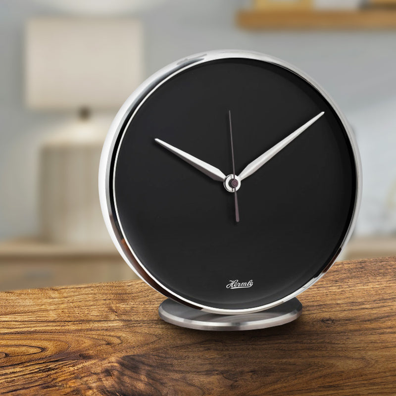 Round desk clock with black dial with metallic hands and rim, sitting on wooden table. 